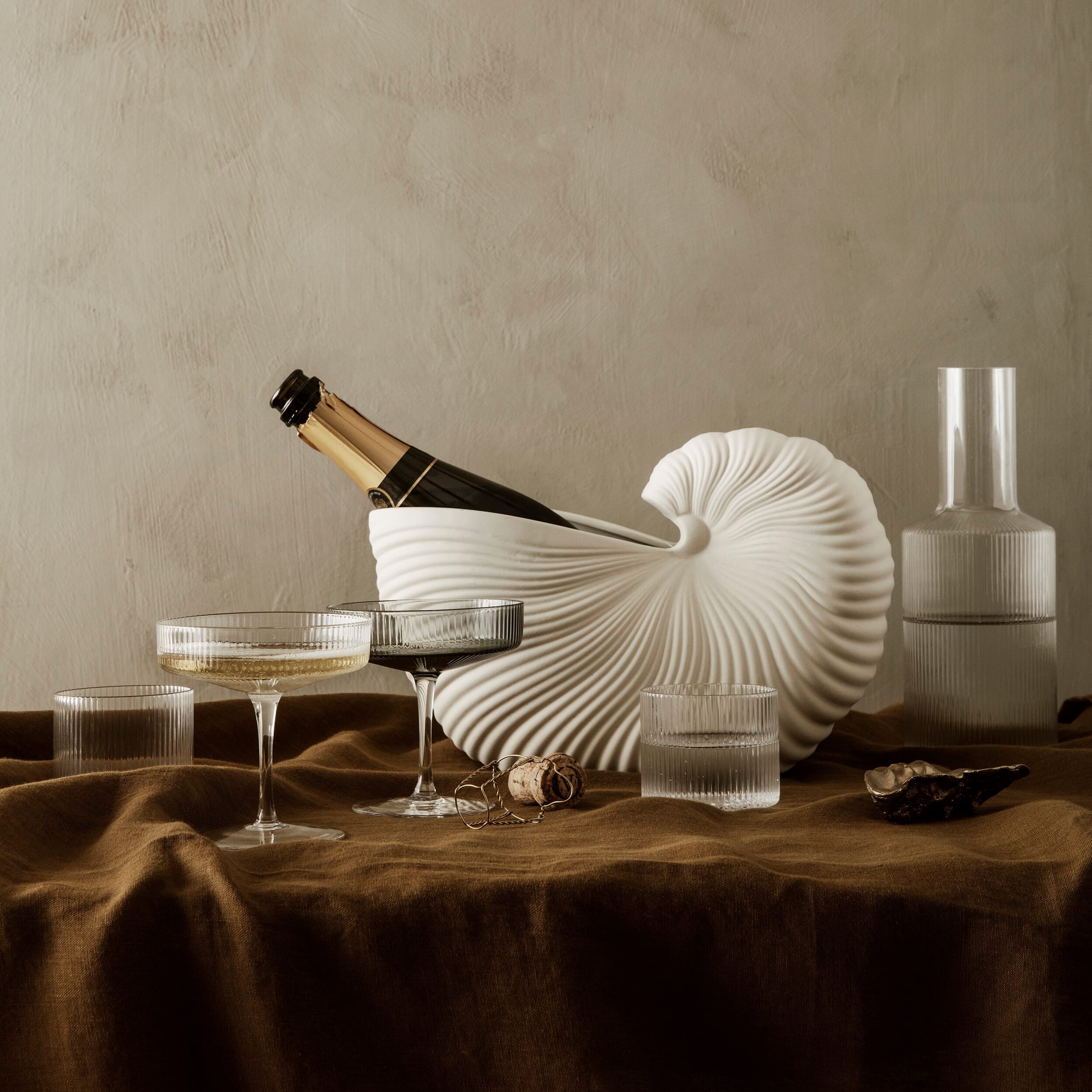 Et stilrent oppsett med elegante glass og en lys, skulpturell vinkjøler formet som en skjellform. En champagneflaske står kjølt i midten, omgitt av serveringsglass og en moderne karaffel på et brunt lintøy som skaper en varm, luksuriøs stemning.