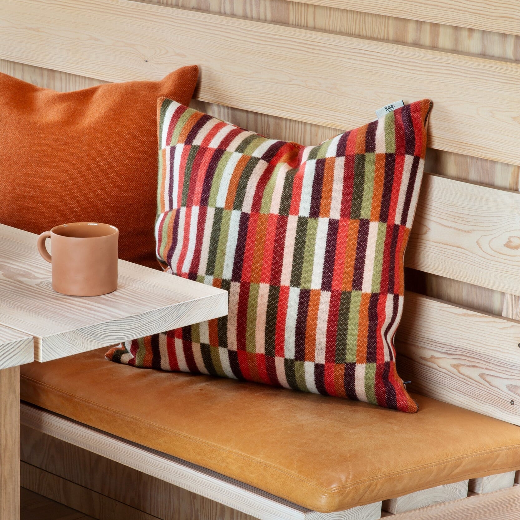 En stor, myk pute med varmt, stripet mønster i rustrøde, grønne og kremfargede toner ligger plassert i en lys trekrok. Ved siden av står en ensfarget terrakottafarget pute og en kopp i samme varme nyanse på et lyst bord – et lunt, hyggelig hjørne.