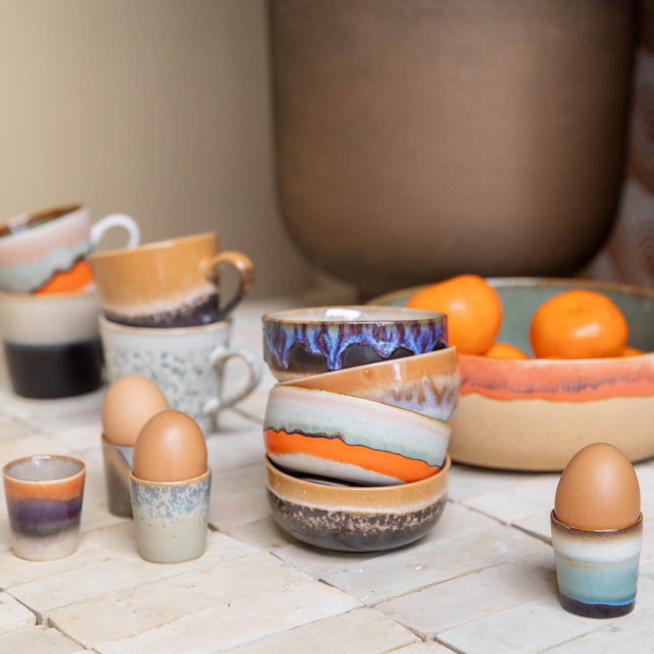 Fargerike keramikkopper, skåler og eggeglass i håndlaget stil, arrangert på kjøkkenbenk med frukt i bakgrunnen.