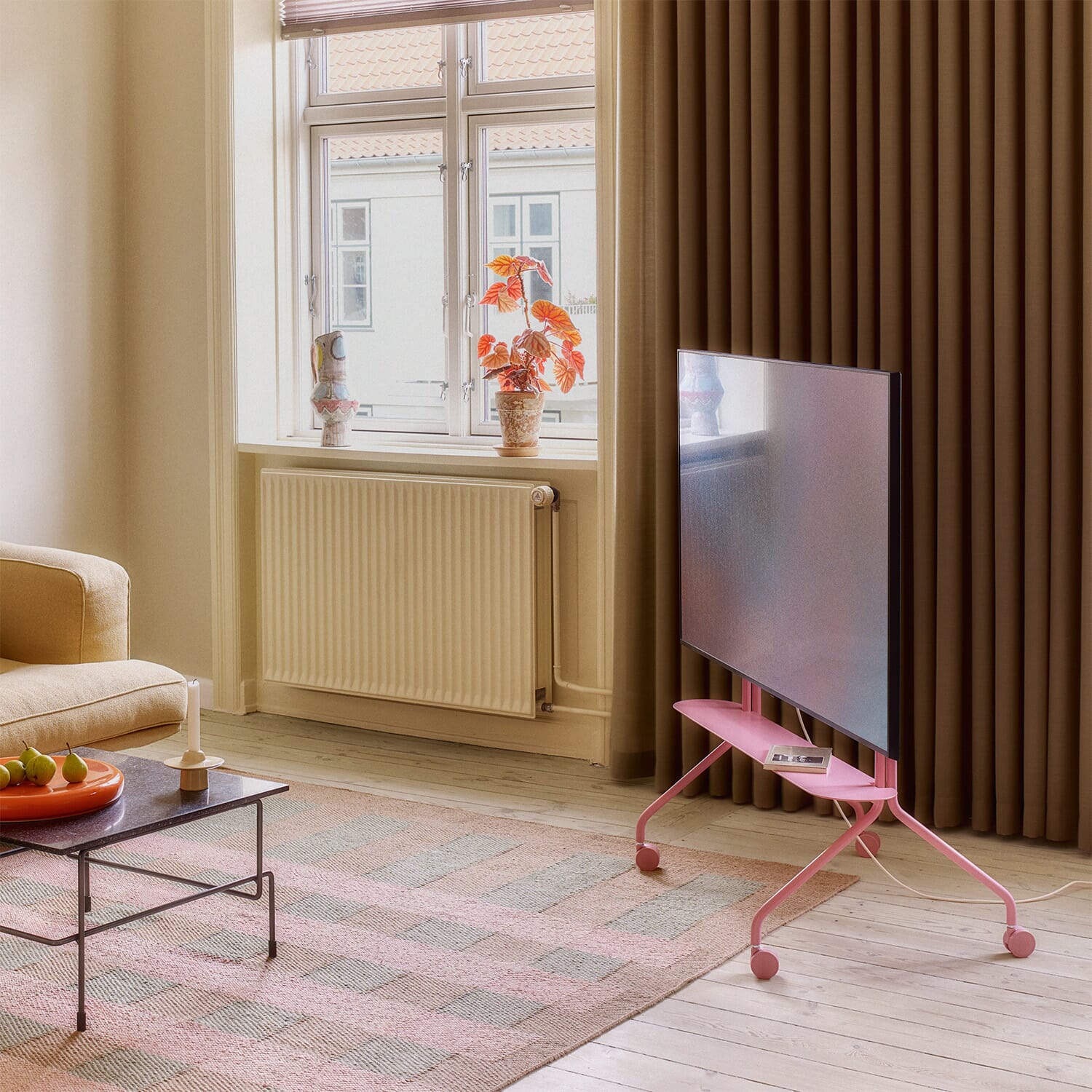 Et lyst og moderne rom med et rosa TV-stativ fra Pedestal på hjul som holder en stor flatskjerm. Under skjermen ligger en bok på den lille hyllen. Myke farger, lyse tregulv, en beige sofa og et vindu med planter skaper en lun og nordisk atmosfære.