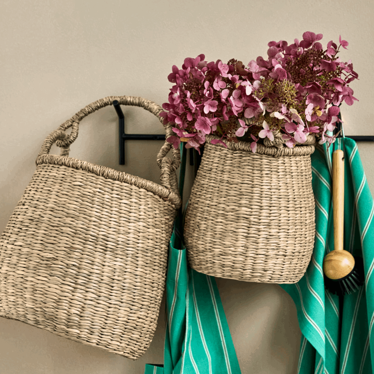 To håndflettede kurver i naturmateriale henger på en svart knaggrekke. Den ene er tom, mens den andre er fylt med rosa hortensiablomster. Under kurvene henger grønne kjøkkenhåndklær og en treskrubb, som skaper en varm og naturlig stemning.