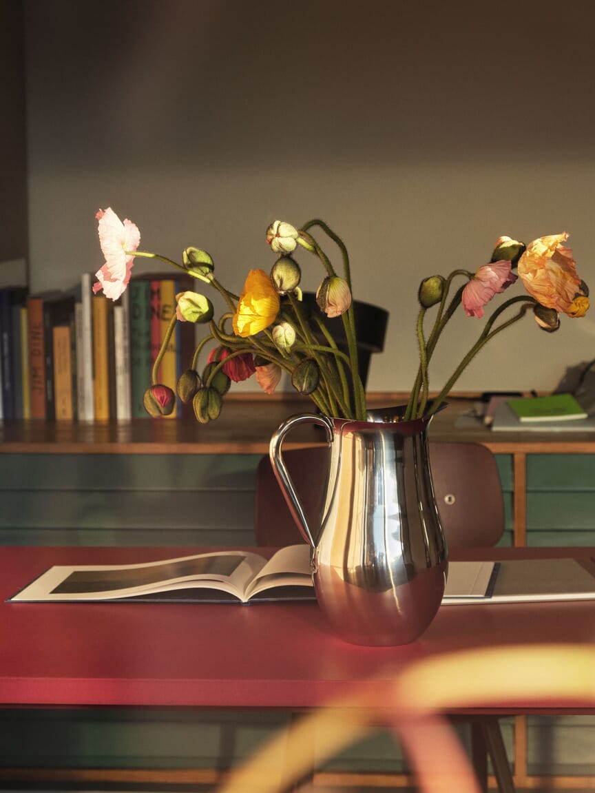 En blank, metallisk kanne står på et rødt bord og brukes som vase for fargerike blomster i mykt ettermiddagslys. Bak sees en bokhylle og en åpen bok, som skaper en varm, rolig og kreativ stemning i rommet.