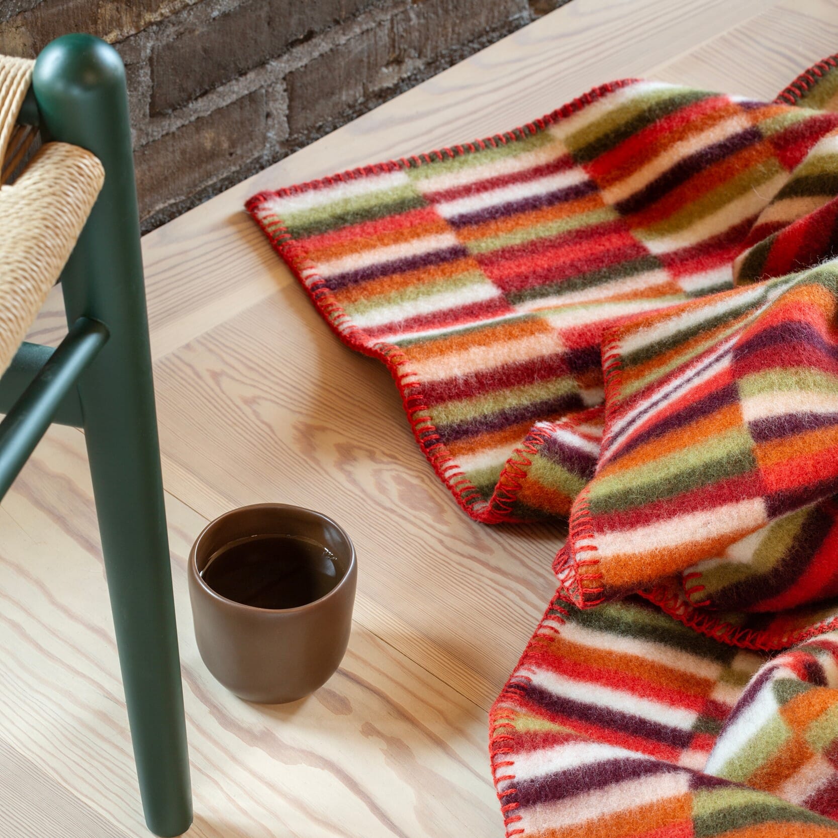 Et fargerikt, rutete pledd i varme røde, grønne og oransje toner ligger mykt over et lyst tregulv. Ved siden av står en enkel brun kopp med varm drikke og et stolbein i grønn finish. Bildet gir en lun, koselig og høstlig stemning.
