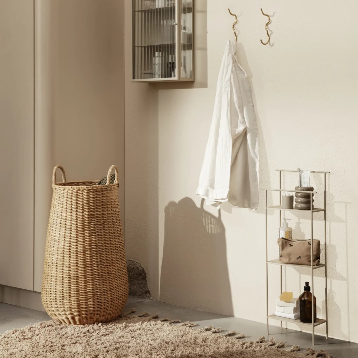 Et lyst og rolig baderom med en høy kurv i naturflett, en beige vegghylle med to kroker og en hvit skjorte, samt et smalt metallstativ med baderomsprodukter. Myke skygger og varme toner skaper et harmonisk uttrykk.