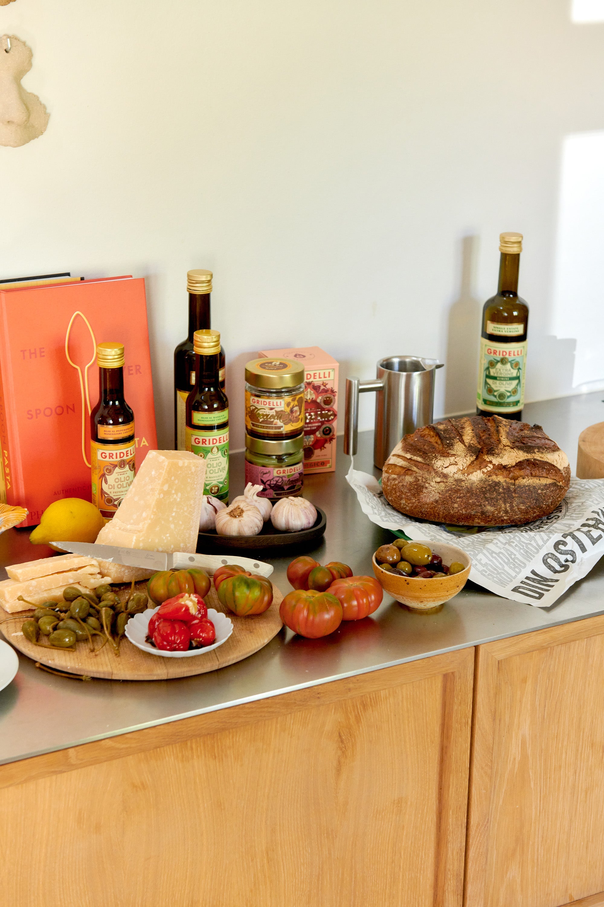 Et kjøkkenbenk-stilleben med delikatesser: flasker olivenolje, syltede grønnsaker, hvitløk, parmesan, tomater og oliven er arrangert ved siden av et nybakt brød. Fargerike råvarer og bøker skaper en varm, rustikk og innbydende matstemning.