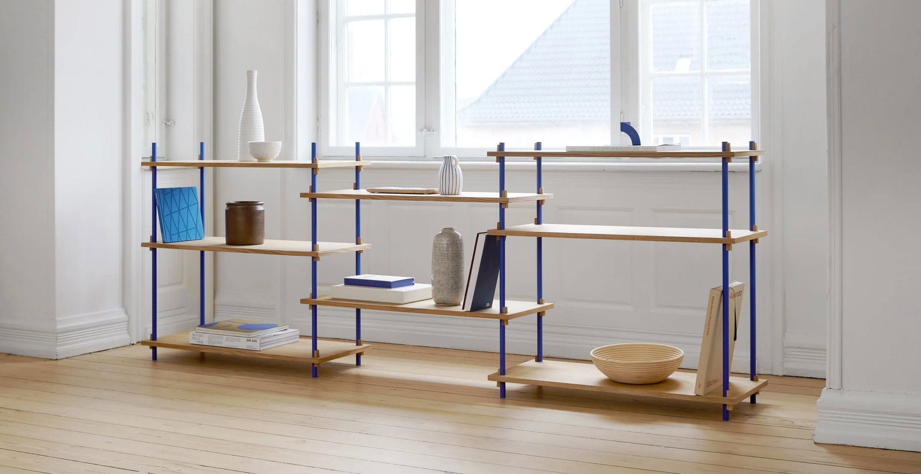 Modulært hyllesystem i lyst tre med blå metallstag, stylet med bøker og keramikk i et lyst skandinavisk rom med store vinduer og tregulv.