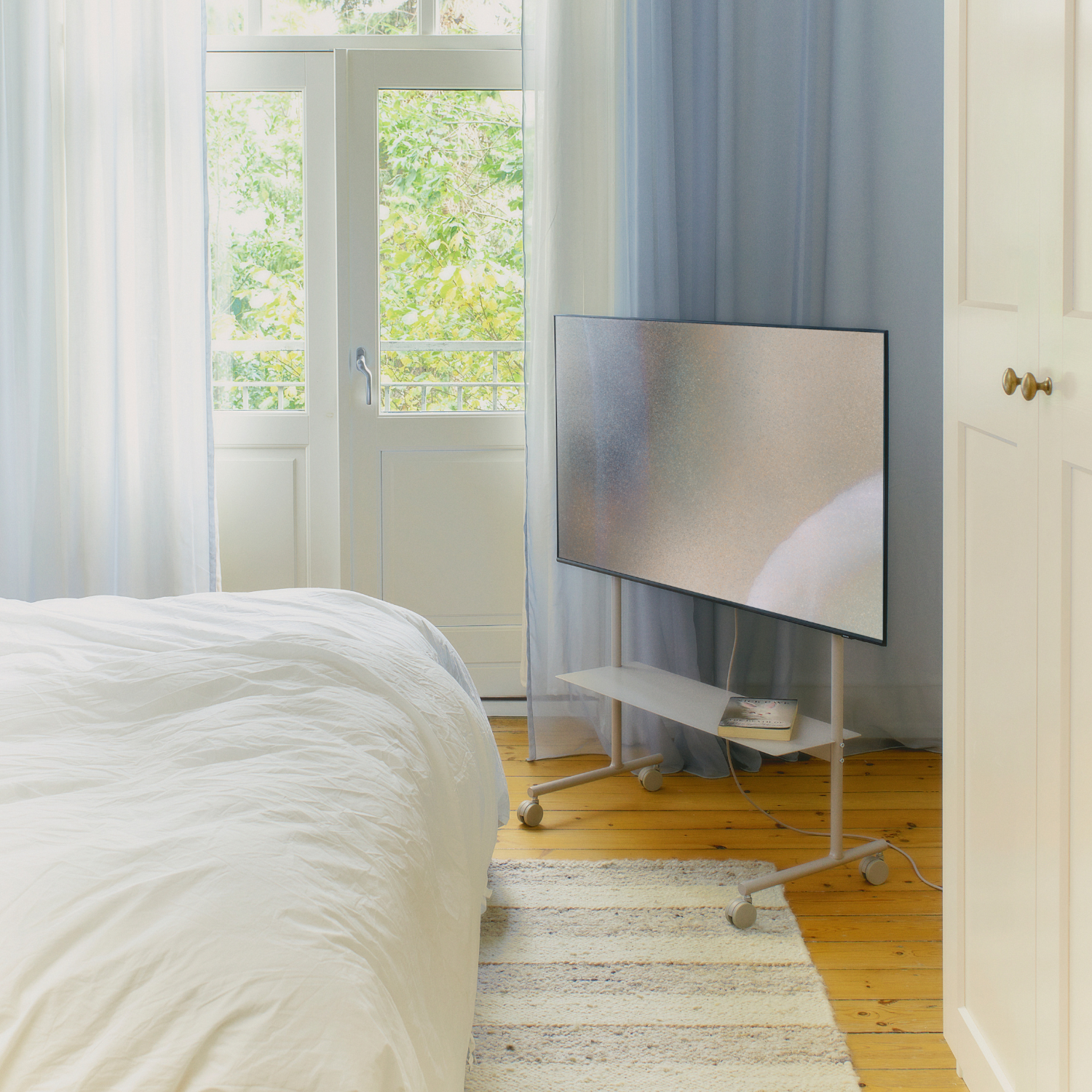 Lys og stilren stue med beige sofa, lavt trebord med bok og potteplante, samt TV på hjul plassert på hvitt tregulv bak delvis åpen glassdør.