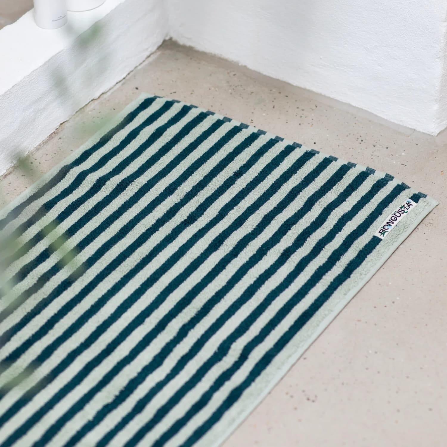 En myk badematte med smale, horisontale striper i mørk grønn og lys mint ligger på et lyst betonggulv. Den rene teksturen og dempede fargene gir et rolig, moderne uttrykk, og et lite merke i hjørnet viser brandnavnet BONGUSTA.