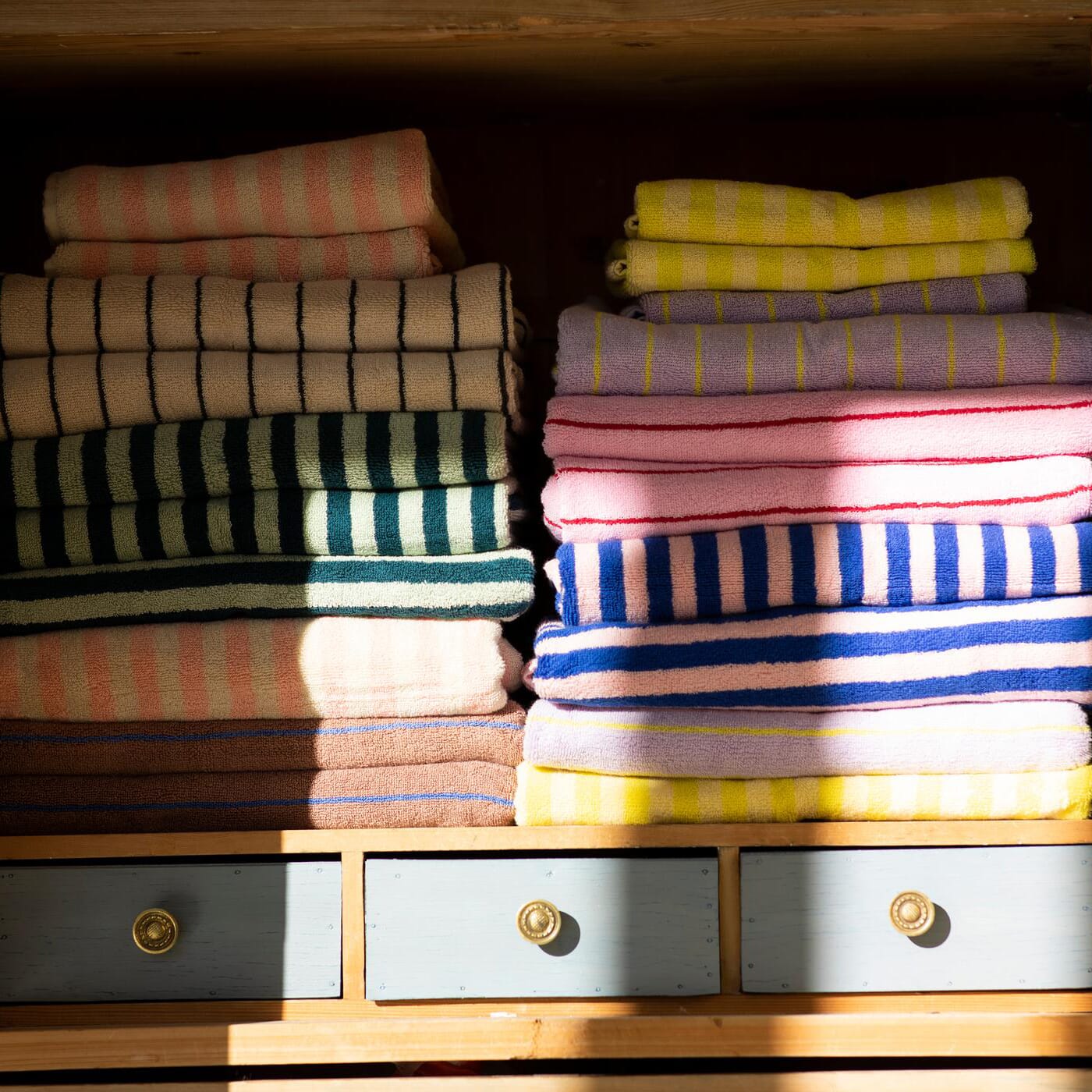 En hylle fylt med pent brettede håndklær i myke beige- og bruntoner, kombinert med lekne pasteller i rosa, blått, gult og lavendel. Stripemønstre gir et livlig uttrykk, mens sollyset skaper varme skygger over teksturen og den lille kommoden under.
