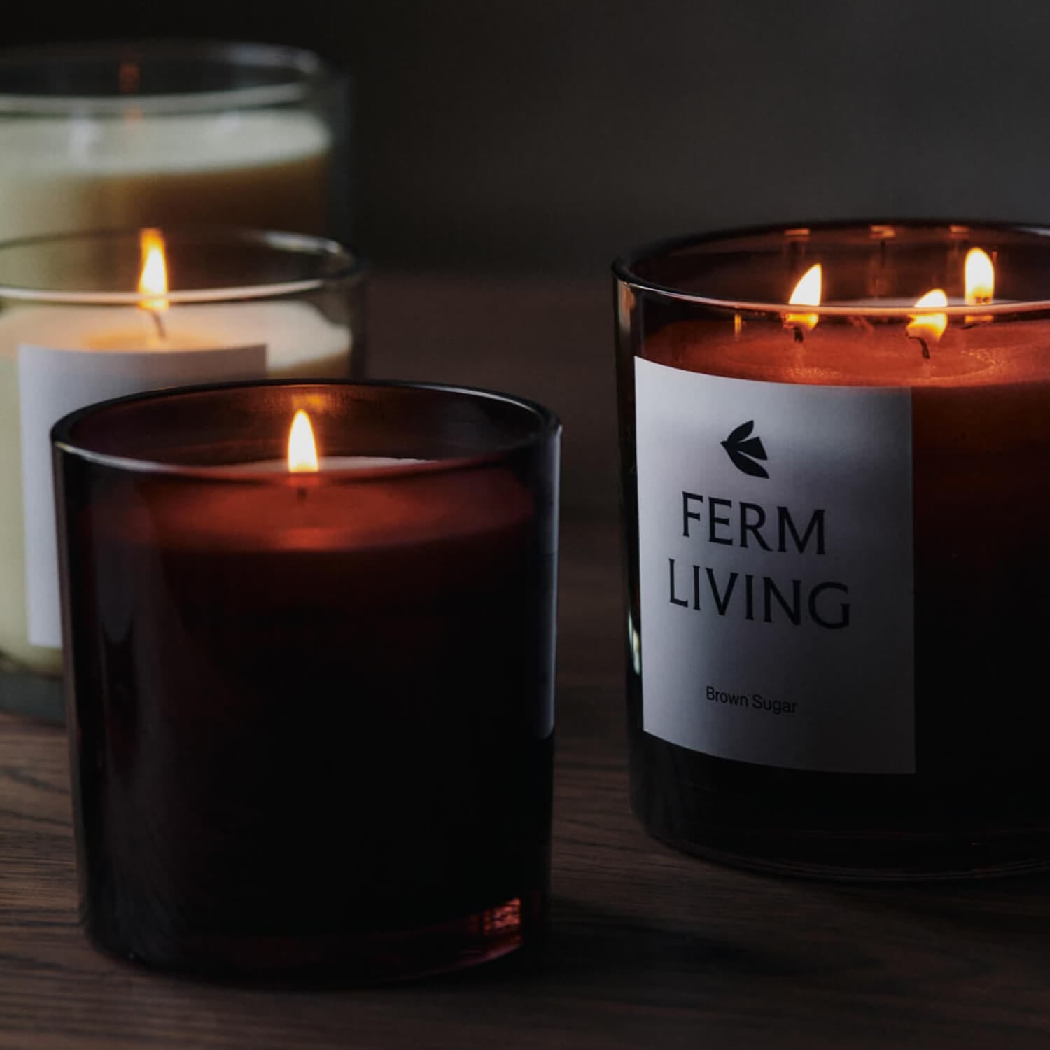 Flere tente duftlys i mørke glass beholdere står samlet på et trebord. Ett av lysene er fra Ferm Living med etiketten “Brown Sugar”. Det varme, myke lyset fra flammene gir en rolig og intim atmosfære i det mørke rommet.