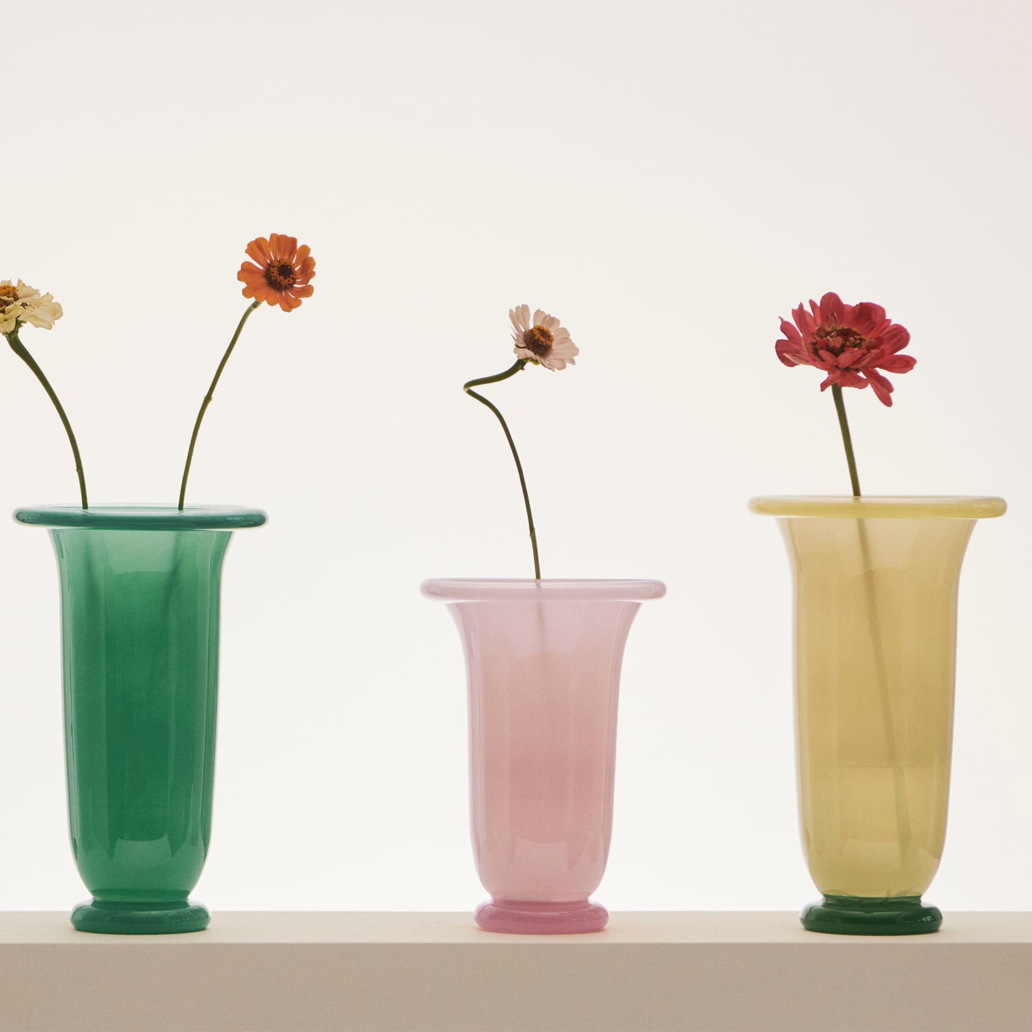 Tre slanke glassvaser i pastellfargene grønn, rosa og gul står på rad, hver med en enkelt blomst med lang stilk. Det rene lyset bak skaper et mykt, minimalistisk uttrykk og fremhever de delikate tonene i vasene.