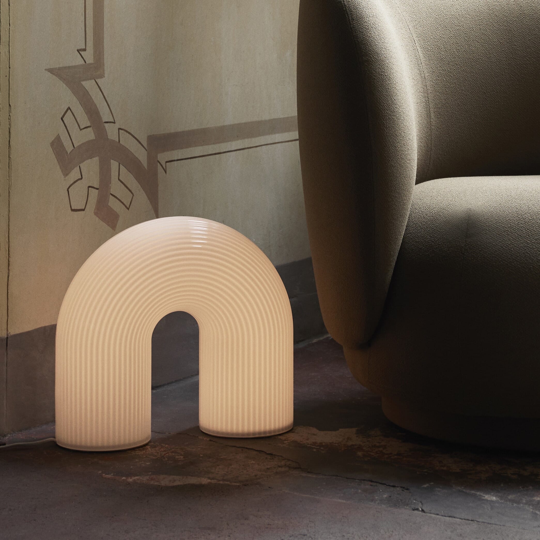 En skulpturell, buet gulvlampe i ribbet, melkefarget glass står på et mørkt gulv ved siden av en stor, beige loungestol. Lampen gir et mykt, varmt lys og fungerer som et dekorativt objekt mot den detaljrike veggen bak.