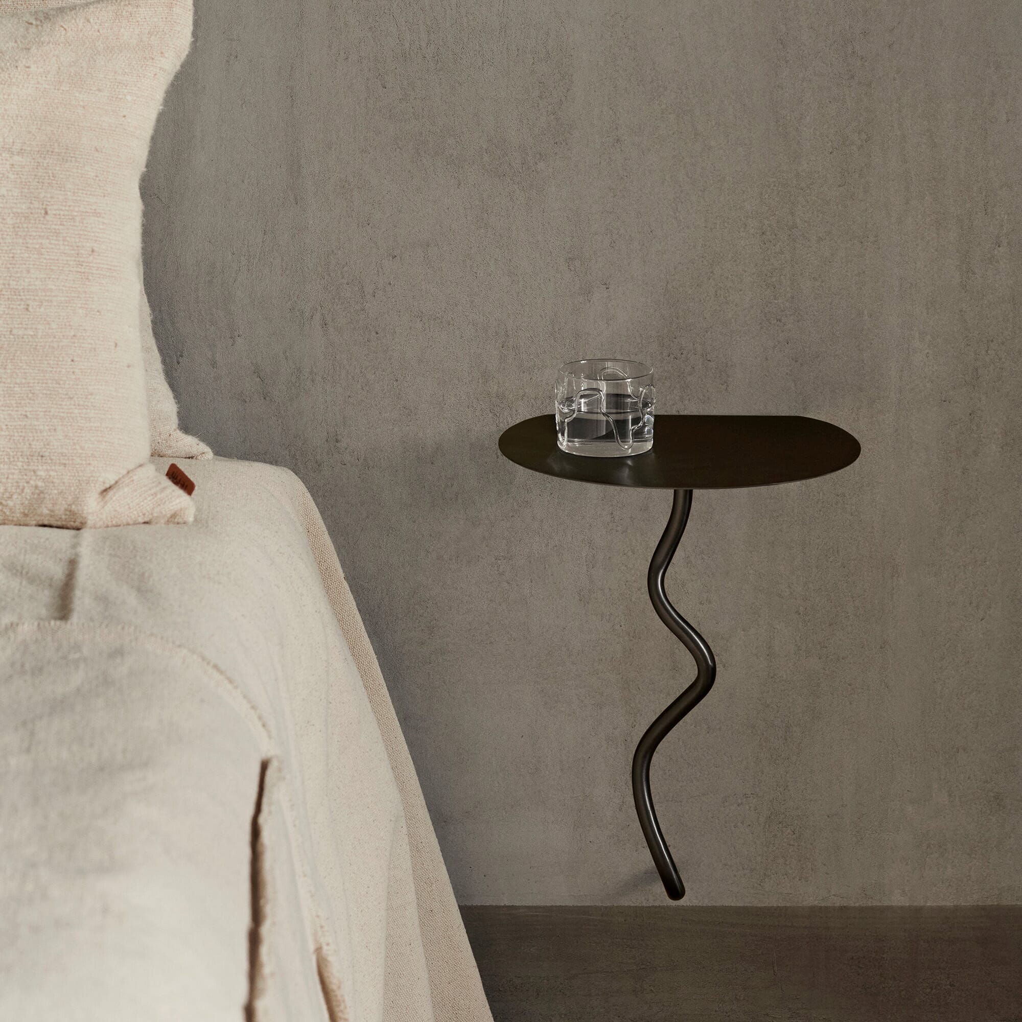 Vegghengt nattbord i mørk metall ved siden av en seng med lyse tekstiler, stylet med et glass vann.