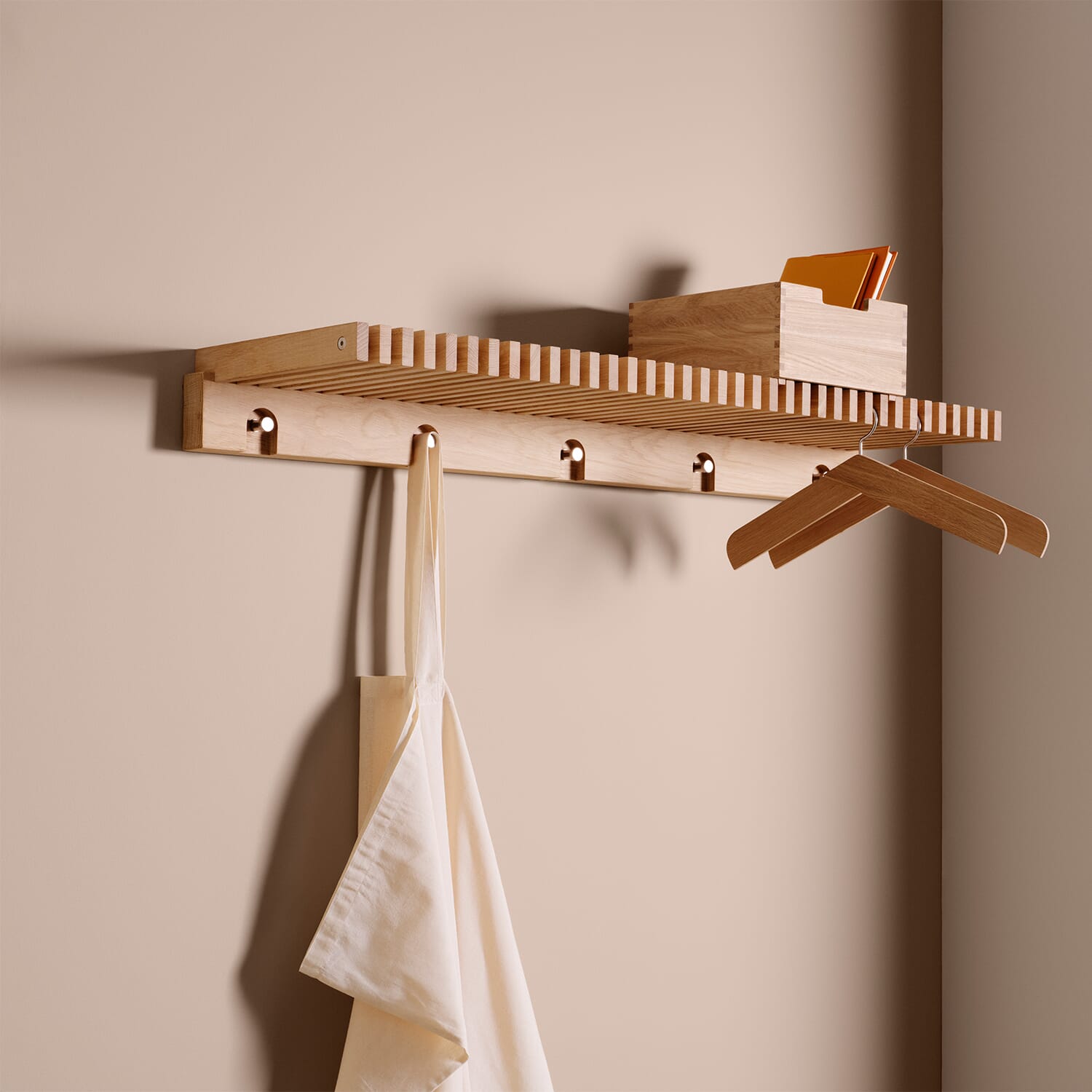 En vegghengt hattehylle i lyst tre med hylleplate og flere knagger under. På hyllen står en liten treboks, og under henger et stoffnett og to trekleshengere. Veggen er malt i en varm beige tone.