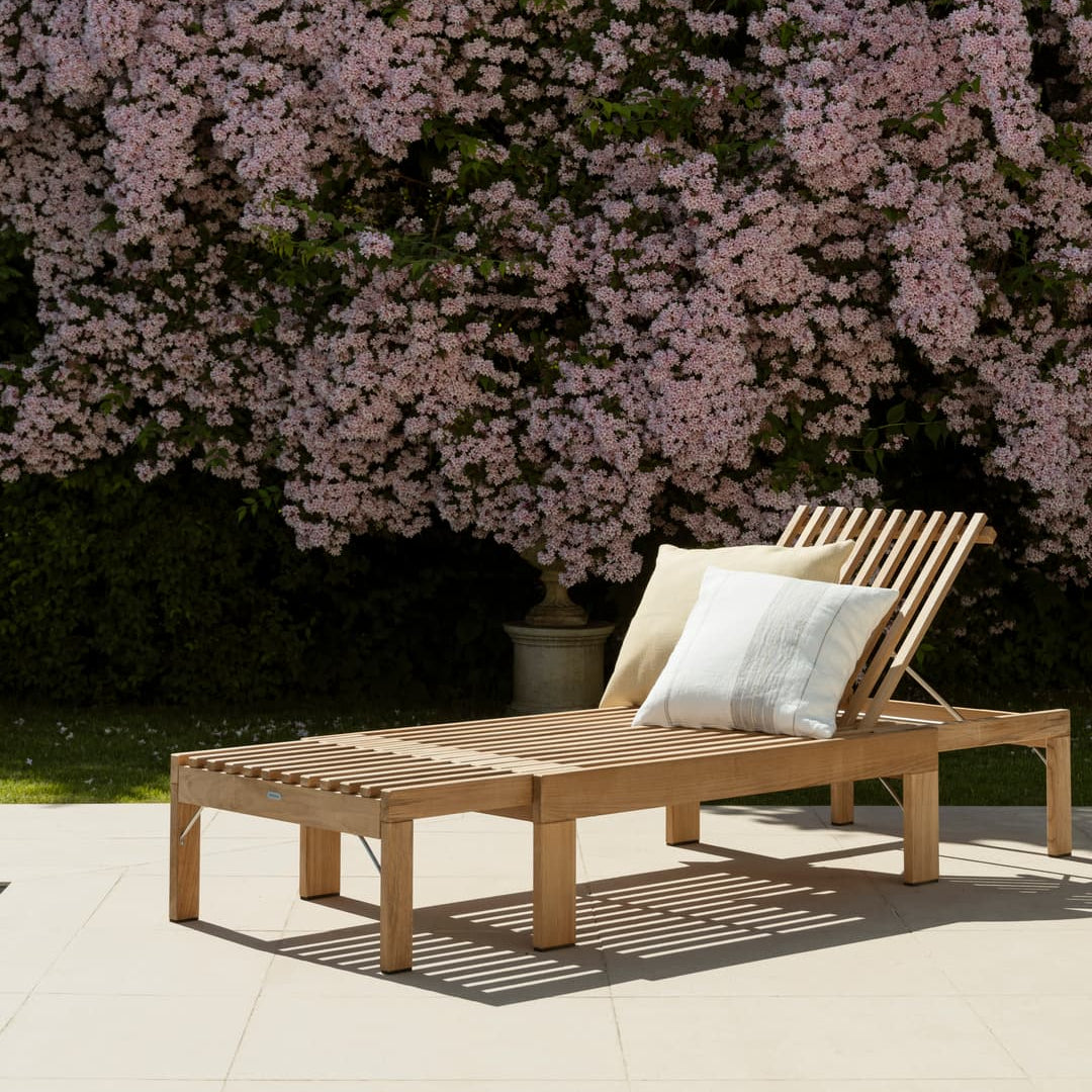 Solseng i teak med justerbar rygg og matchende fotskammel på terrasse ved moderne hytte, omgitt av gress og furutrær i sommerlys.