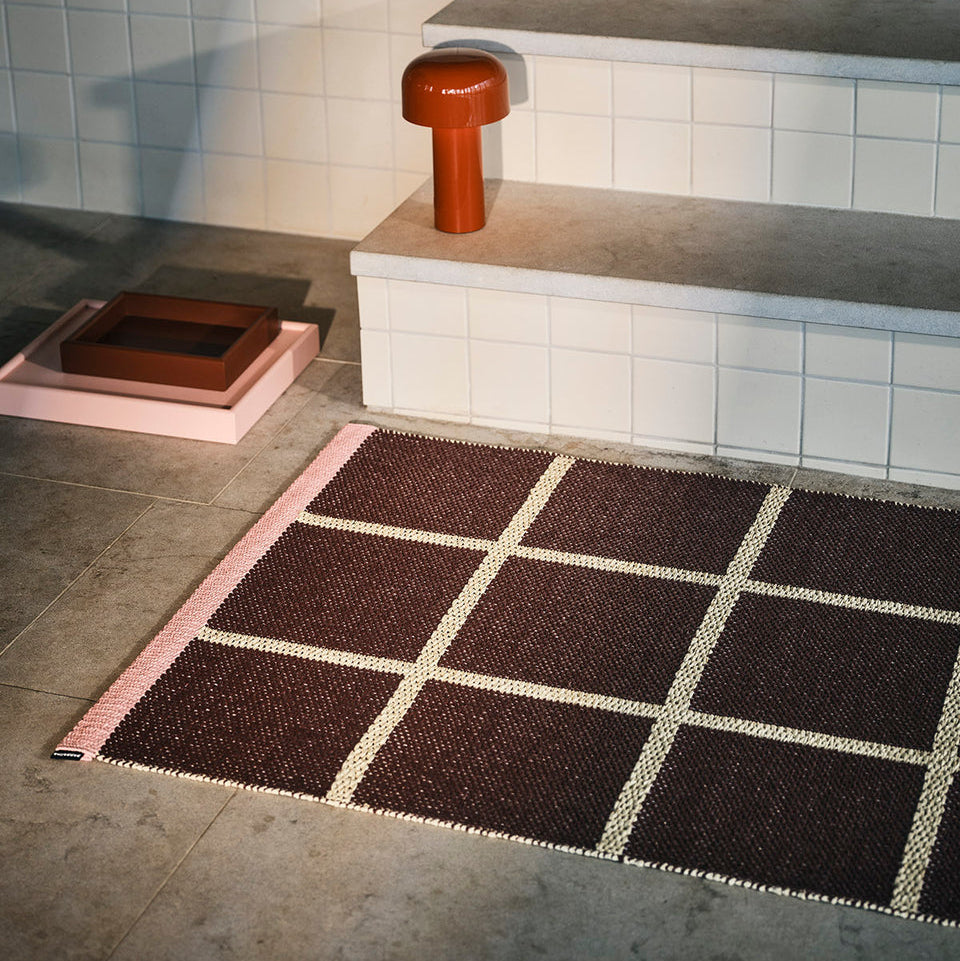 Rektangulært håndvevd teppe i brun ull med rutemønster i krem og rosa kant, plassert på flislagt gulv ved trapp i moderne interiør.