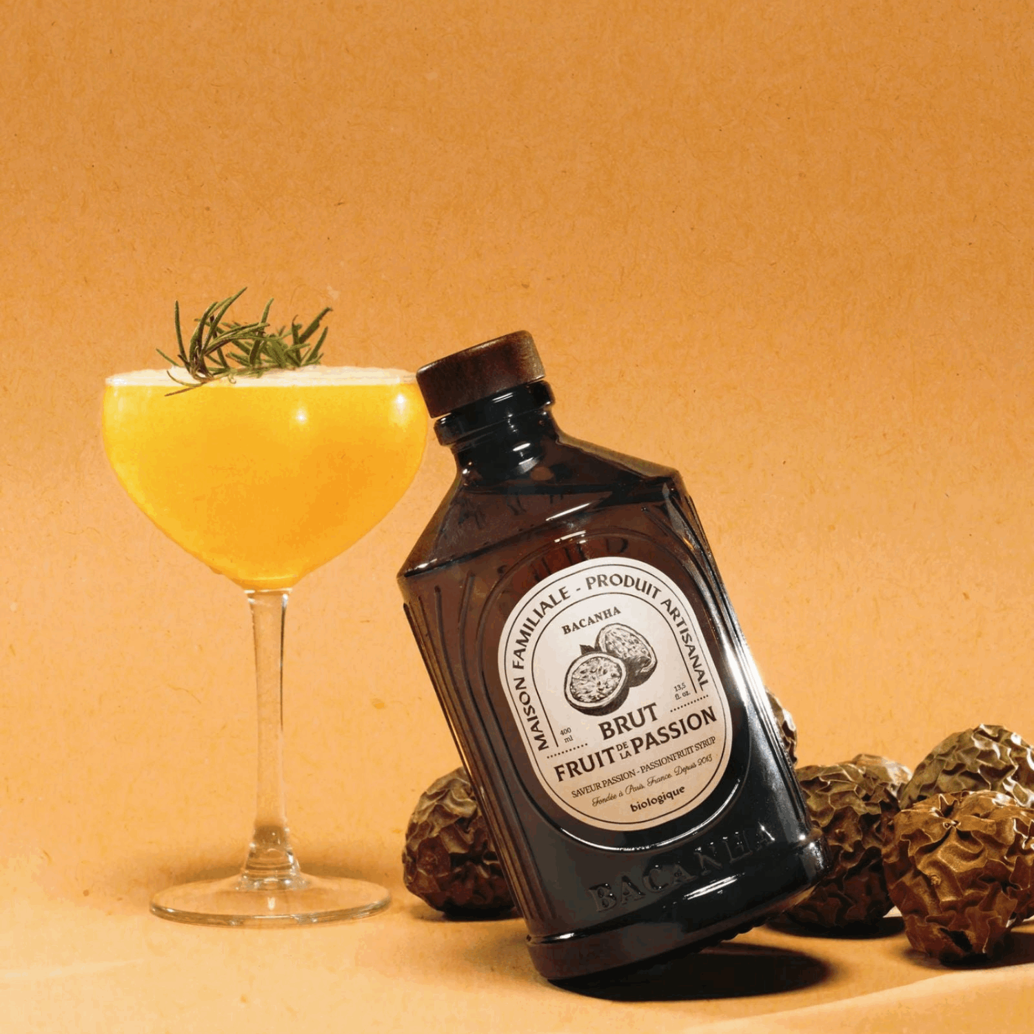 Et elegant cocktailglass fylt med gul drink ved siden av en mørk glassflaske med etiketten Brut Fruit de Passion. Rundt flasken ligger tørkede pasjonsfrukter. Den varme, beige bakgrunnen gir et luksuriøst og innbydende uttrykk.