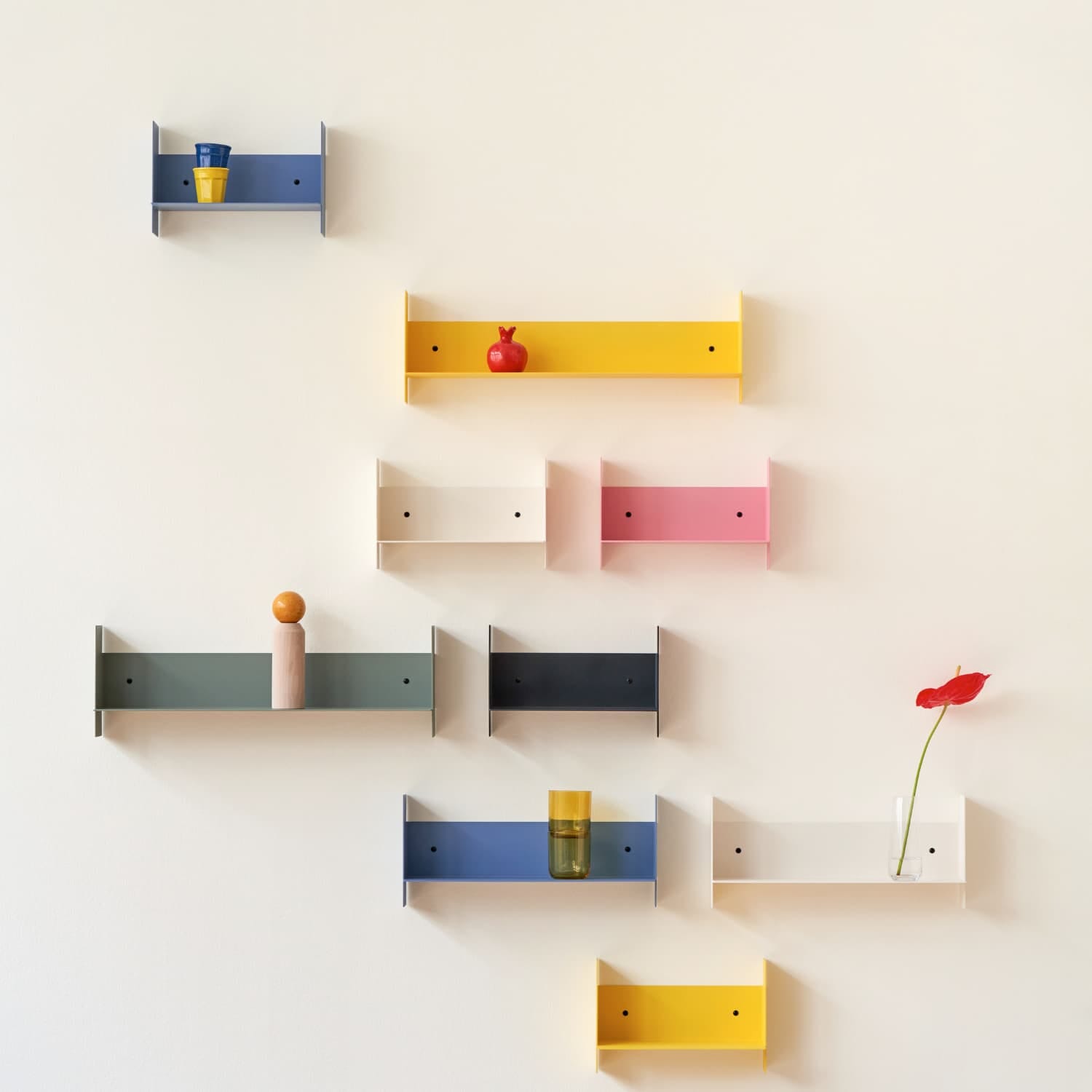 Vegghengte hyller i ulike farger som gul, blå, rosa, grønn og hvit, arrangert dekorativt på lys vegg med små vaser og dekorobjekter.