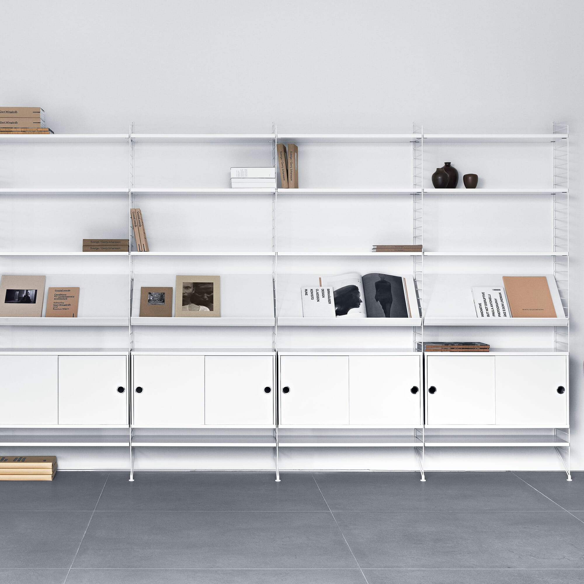 Et stort hvitt String-hyllesystem dekker veggen, med åpne hyller øverst og skapmoduler nederst. Hyllene er stylet med bøker, magasiner og keramikk. Rommet har lyse vegger og grå gulvfliser i en ren, skandinavisk stil.