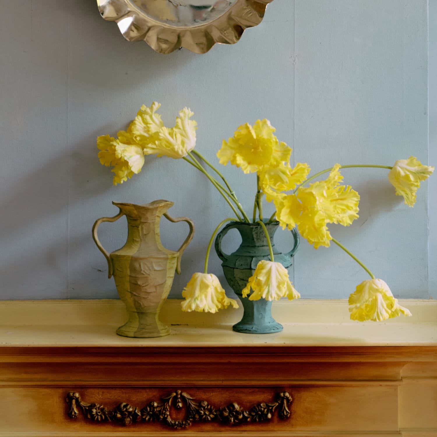 To dekorative vaser står på en utsmykket peishylle, fylt med store gule blomster som bøyer seg mykt utover. Det blå veggpanelet og de varme tretonene skaper et harmonisk og kunstnerisk uttrykk i rommet.