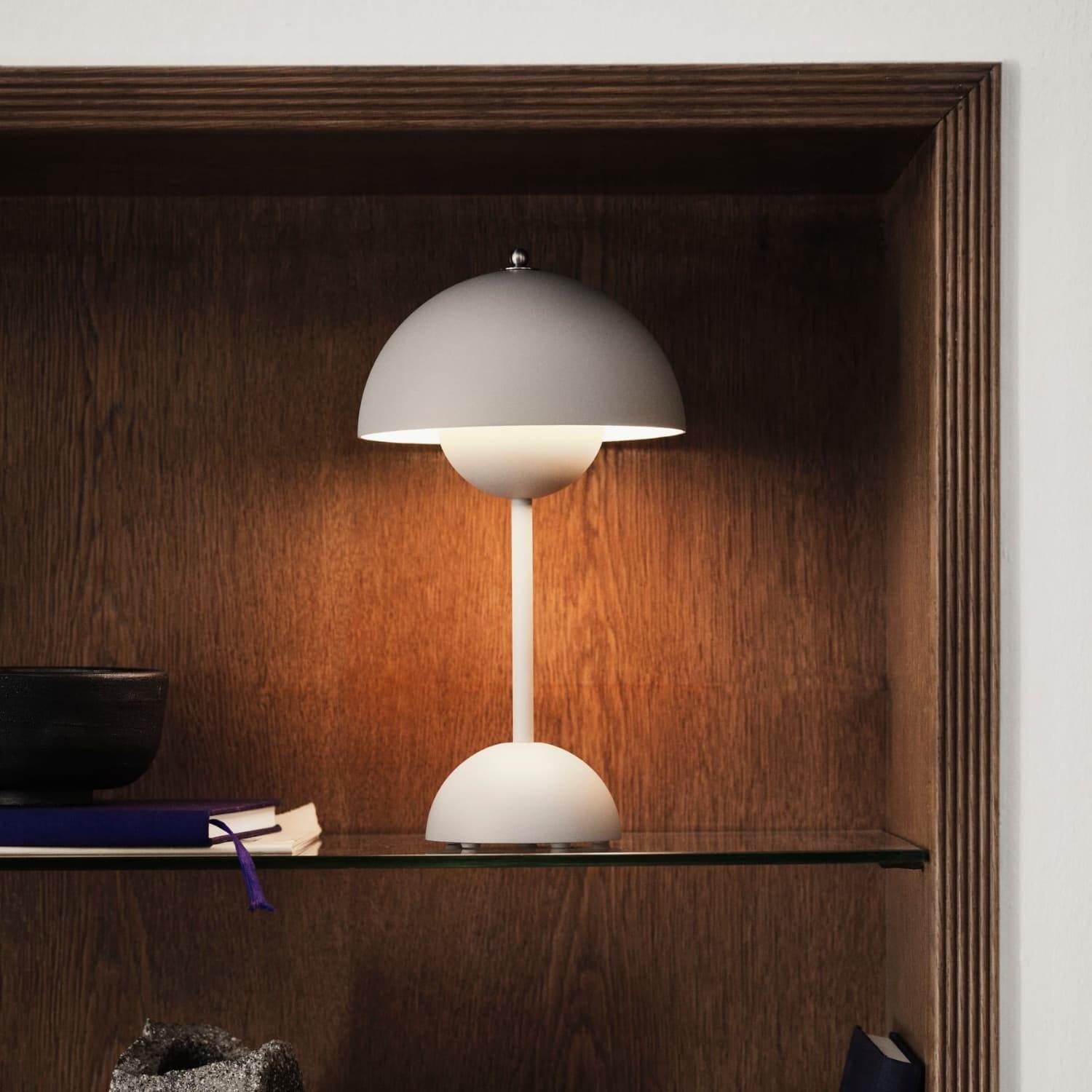 En liten, elegant bordlampe i hvitt står på en glasshylle inne i en nisje av mørkt tre. Det myke lyset skaper en lun glød rundt lampen. Ved siden av ligger en bok med lilla bokmerke og keramikkobjekter som gir et rolig og stilrent uttrykk.