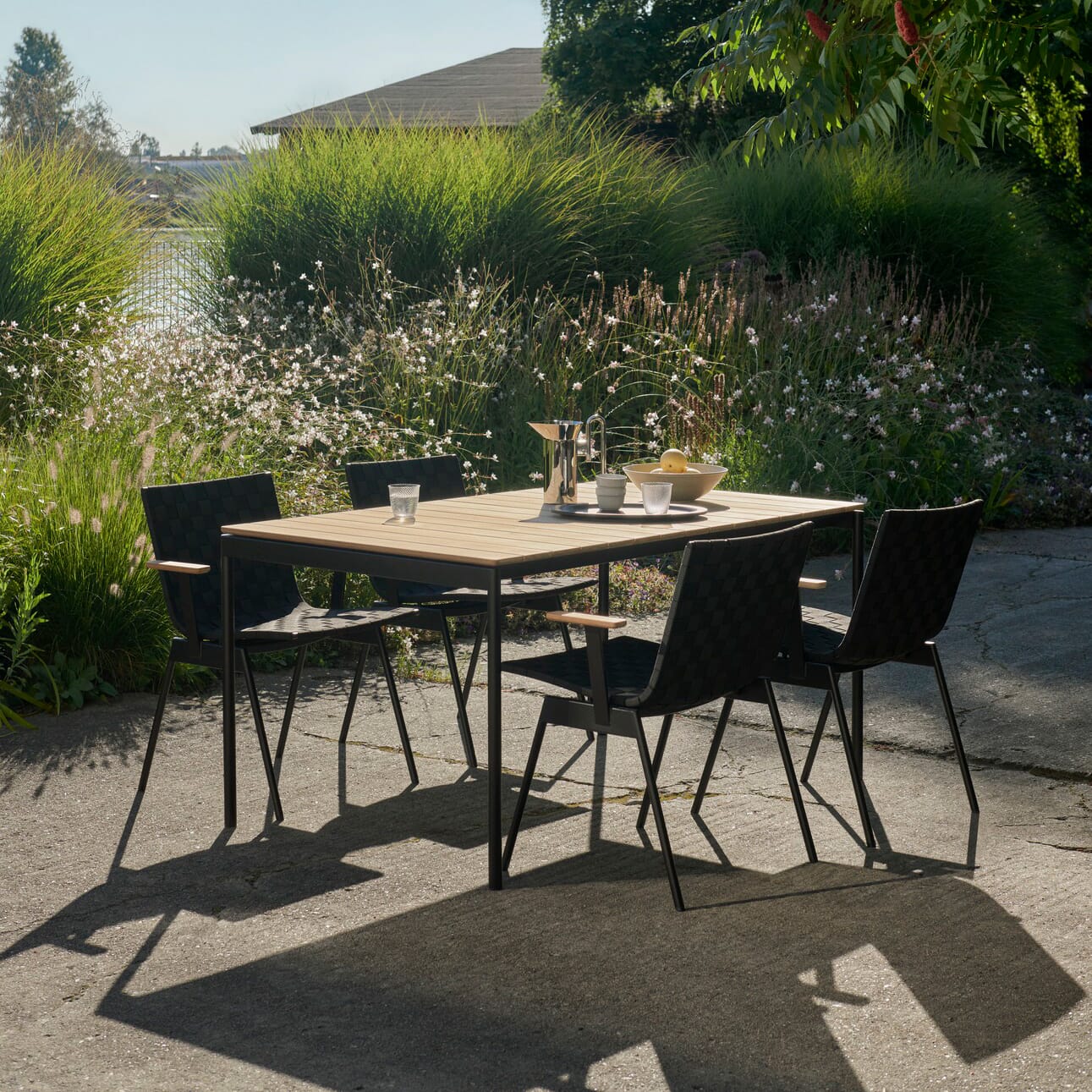 Ville utebord med sort ramme og teakbordplate med oppdekking på en uteplass i grønne omgivelser med stoler rundt.