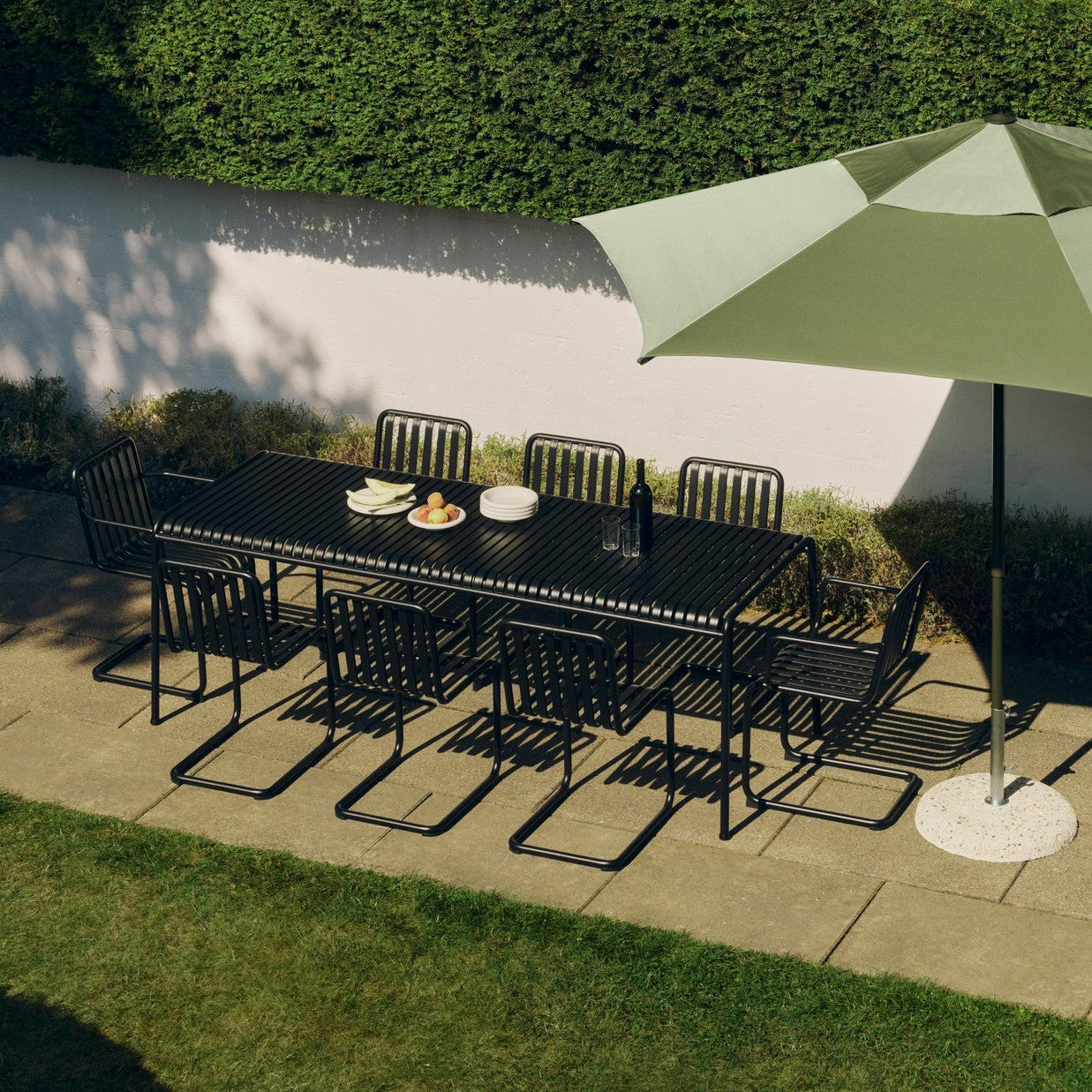 HAY Palissade spisegruppe med bord og stoler på en hellebelagt platting sammen med en grønn parasoll fra HAY