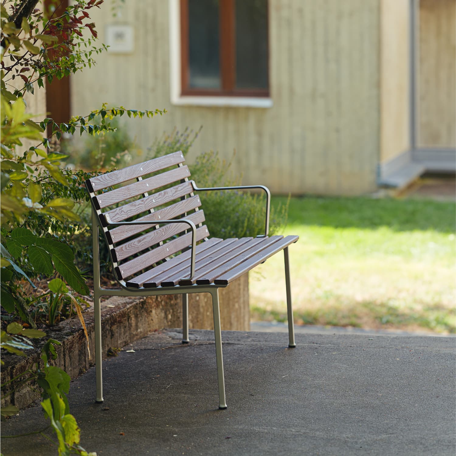 Traverse benk med armlener på en terrasseplatting av betong inntil et bedd med grønne vekster