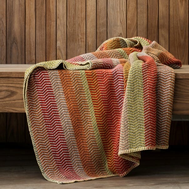 FRI summer red pledd i norsk ull fra Røros Tweed, med myk sikksakk og varme fargetoner, lagt over en trebenk.