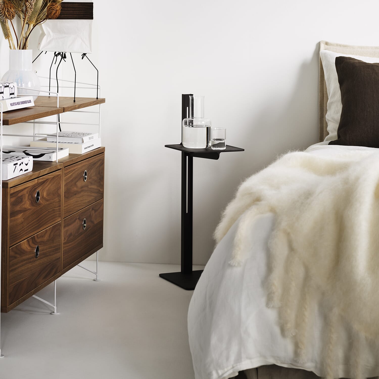 Museum sidebord i svart aluminium ved siden av en seng og en stringhylle med hvite gavler og valnøtt-hyller.