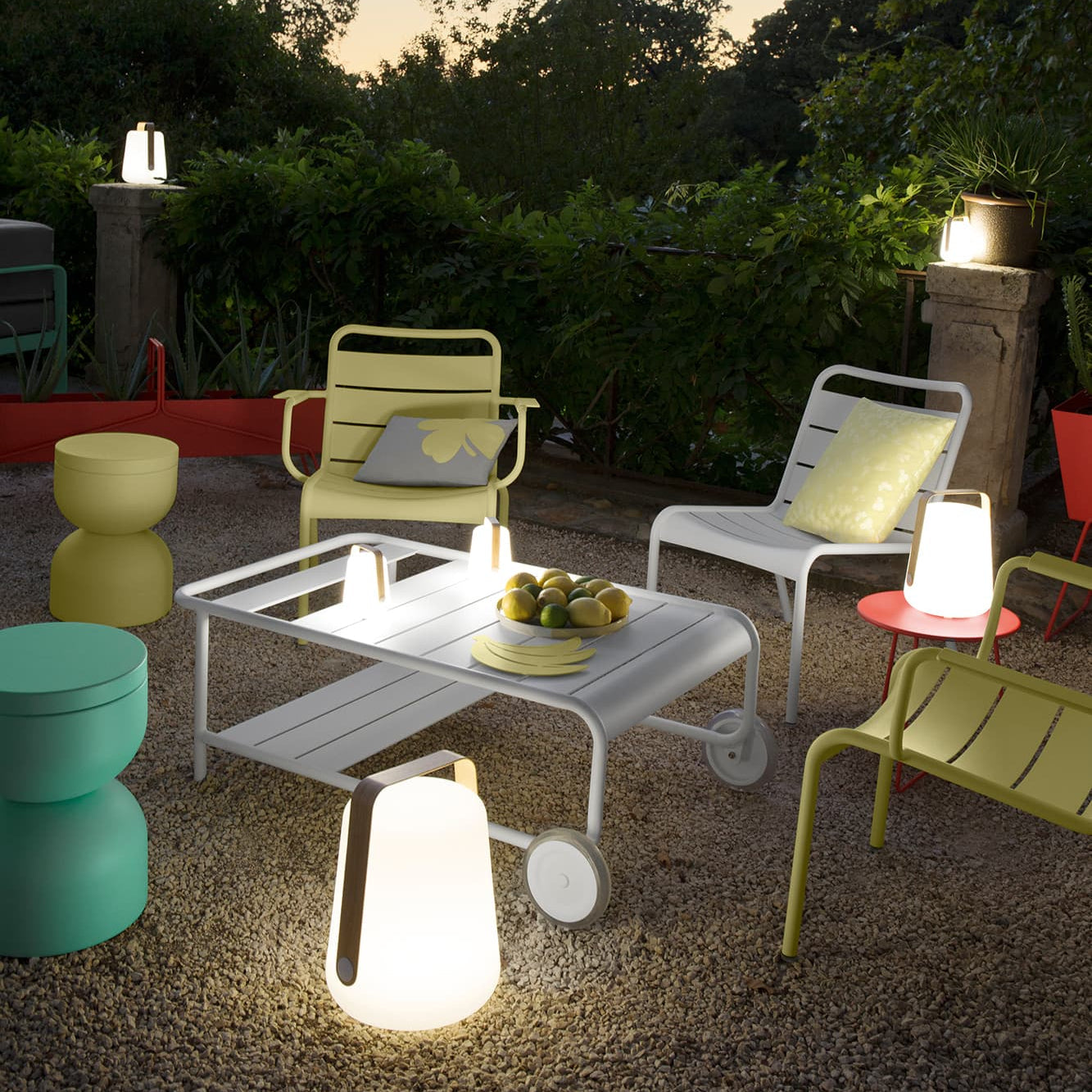 Utendørsmiljø med teakbenk, fargerike puter og små sidebord i metall, dekorert med planter og keramikk – sommerlig terrasseinnredning.