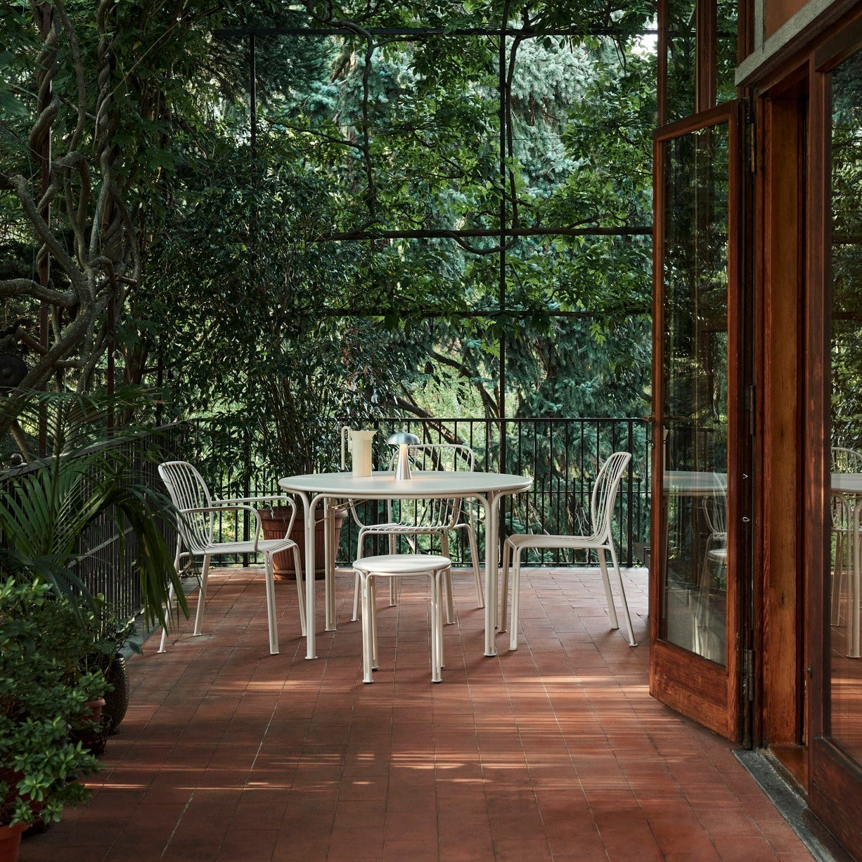 Lyst utemiljø med rundt bord og stoler i metall, plassert på flislagt terrasse omgitt av frodig grønt og store glassflater som slipper naturen inn.