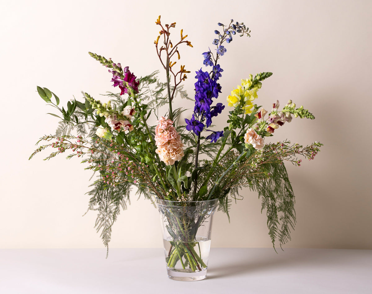 Tre slanke glassvaser i pastellfargene grønn, rosa og gul står på rad, hver med en enkelt blomst med lang stilk. Det rene lyset bak skaper et mykt, minimalistisk uttrykk og fremhever de delikate tonene i vasene.