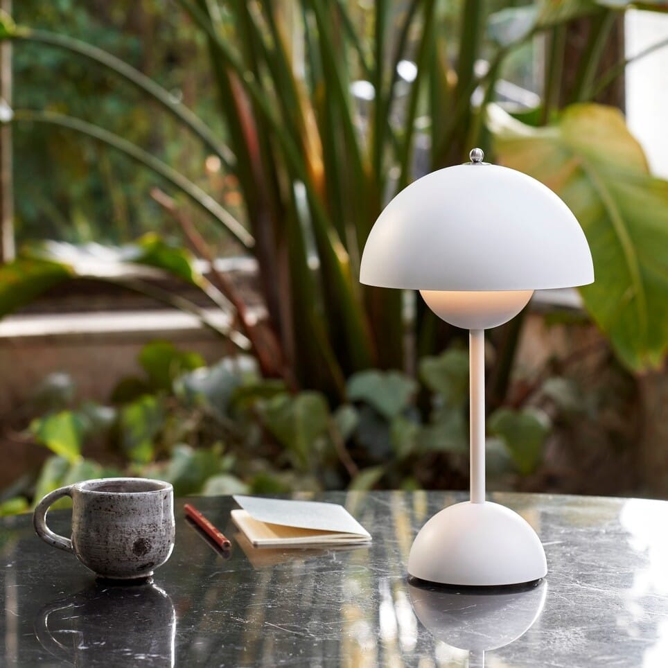 Oppladbar bordlampe i hvitt på et bord ute i hagen, omgitt av grønn planter.