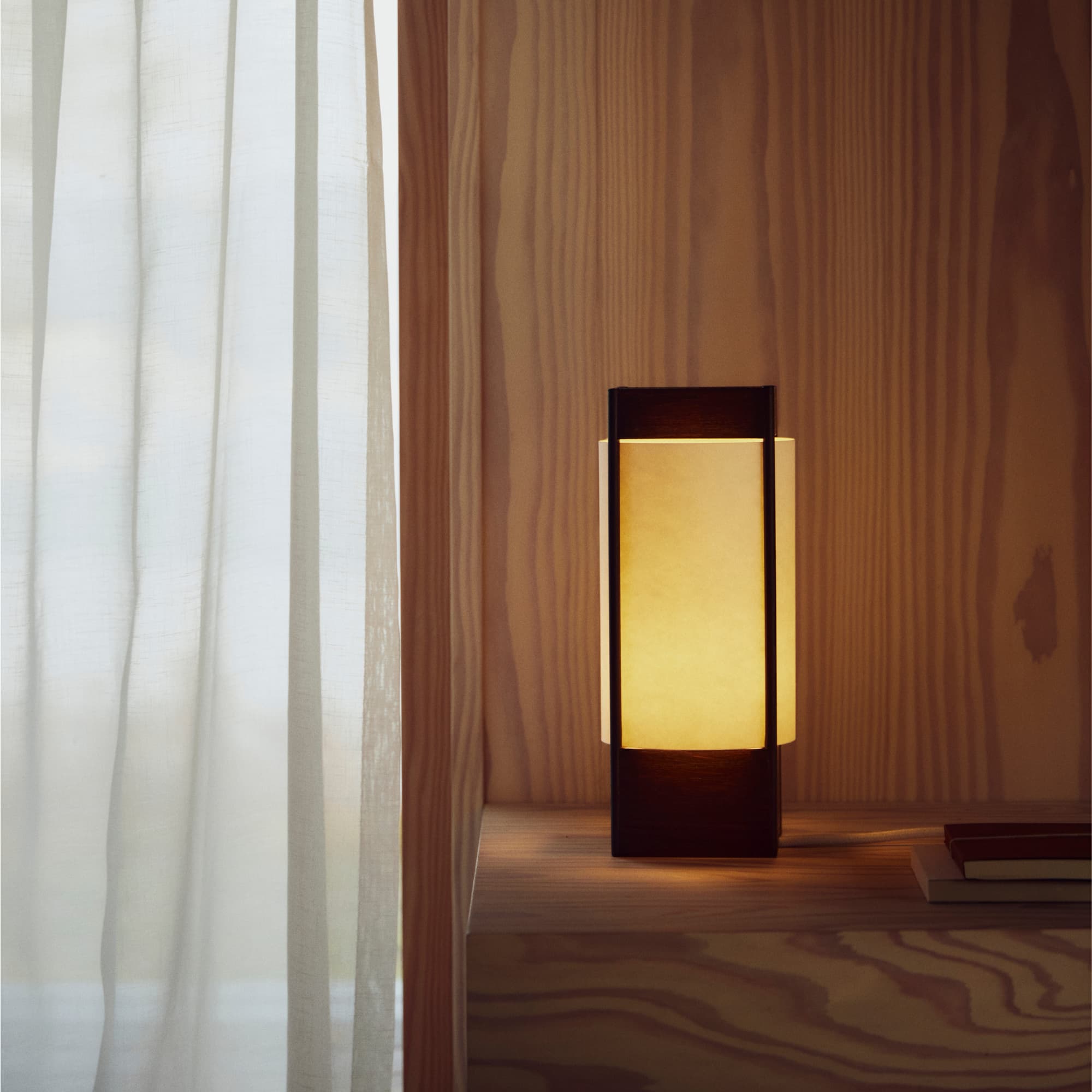 En bordlampe med hvit, halvkuleformet skjerm og base i rustrød marmor står på et skap i lys eik. Ved siden av ligger en bok og en liten skål i lyst tre. Lampen gir et mykt, varmt lys i et minimalistisk rom med lyse vegger.